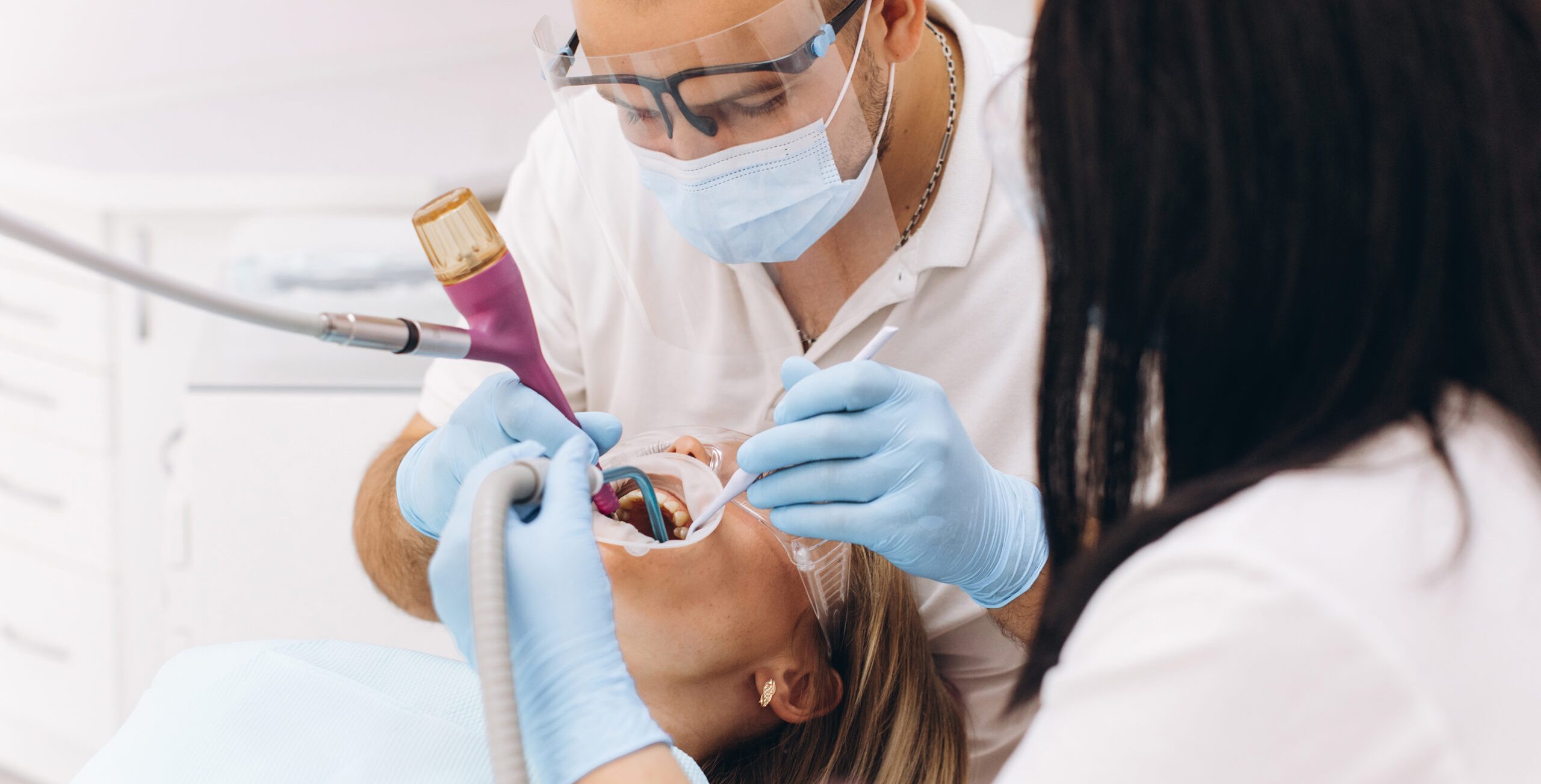 A dentist in a mask and goggles treats a woman's teeth.